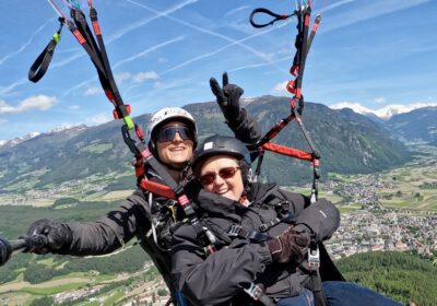 Zwei Personen hintereinander in einem Gurtsitz über der Erde. Im Hintergrund sind Berge. Beide tragen Helme.