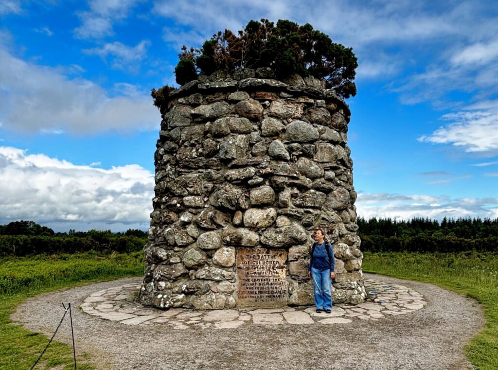 Denkmal der Schlacht von Culloden. Ein runder Turm aus aufgeschichteten Steinen. Davor eine Frau in Jeans und blauer Jacke.